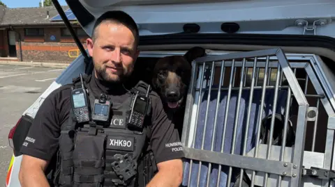 Kevin Shoesmith/BBC A police officer next to a cage, in the back of a police car, containing a brown and black Belgian Malinois
