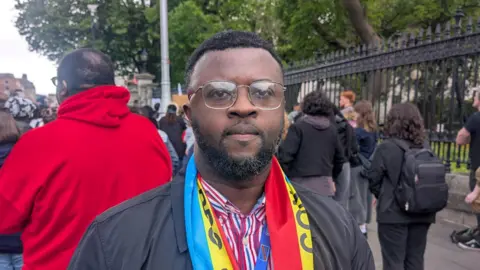 PA Media A antheral   stands successful  a achromatic  overgarment  and bluish  and achromatic  striped shirt. He has abbreviated  achromatic  hairsbreadth  and a abbreviated  achromatic  beard. He wears a yellow, bluish  and reddish  scarf that reads 'Congo' connected  it. 