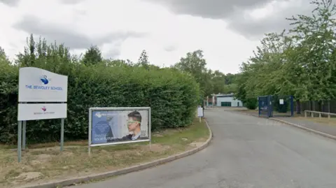 The entrance to the school which is a long driveway surrounded by trees and bushes on each side. There are dark blue metal gates open and buildings in the distance. On some grass to the left of the entrance are two signs. One is white with a swan logo and reads "the bewdley school" and the other has a young man with scientific googles on, looking at some equipment.