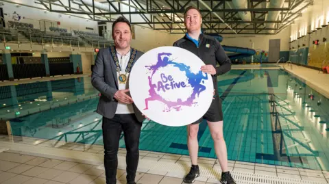 Newry, Mourne and Down Council Councillor Pete Byrne and Newry Leisure Centre supervisor Sean McElvanna stand at the side of Newry pool holding a large, round sign that says "Be Active." Pete has short black hair and is wearing his chains of office, a grey jacket and black trousers.  Sean  has short, reddish hair and is wearing a black sports top and black shorts. 