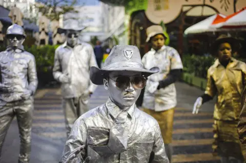 Kim Ludbrook/EPA Street artists painted in gold and silver stand motionless as they wait for donations from passers-by and shoppers at the Rosebank Shopping Mall on 17 June 2023.