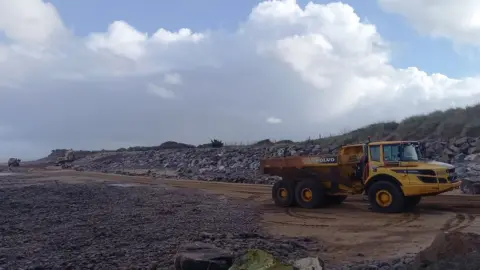 BBC A yellow dumper truck delivering large granite boulders on a sandy beach on a sunny day