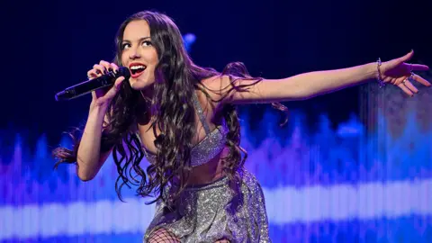 Getty Images A young woman holds a microphone to her mouth and leans forward slightly, causing her long black hair to hang down in front of her. Her other arm is outstretched, as if she's trying to encourage the crowd to sing along with her. Behind her an elaborate arc of stage lights glows bright blue.