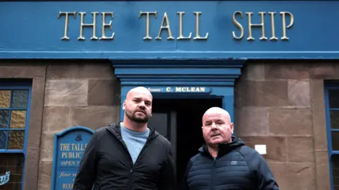 BBC James Lacey and his son standing outside the Tall Ship pub. The pub is brick and painted in blue. Father and son both have bald heads and are wearing jackets. The younger man has a beard.