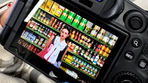 A girl with a bold outfit stood in front of shelves inside a corner shop.