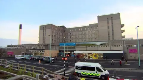 BBC Derriford Hospital. It is a large concrete building with a road running in front of it which has an ambulance driving along it.
