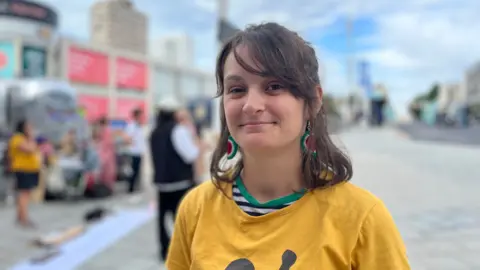 Cat a mother of two wearing a yellow t shirt with brown shoulder length hair and water melon earrings, behind her is a crowd with placards outside Churchill Square shopping centre