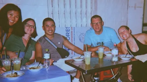 Ike Robin Ike and his adoptive family are all sitting on smiling around a table. Ike is between his mum and dad. His sister and girlfriend are also smiling to the camera. On the table there are bowls of food and plastic cups with green straws. 