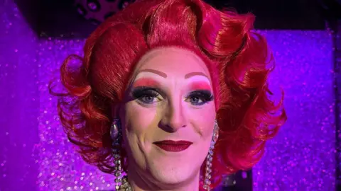 A drag performer in stage make up and a flamboyant red wig curled away from her face. She is wearing long diamante earrings and standing in front of a sparkly backdrop.