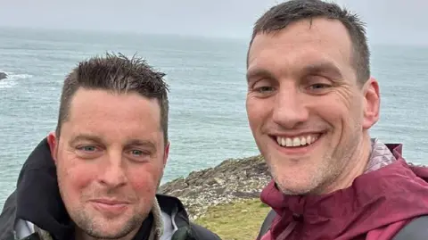 Maxwell Family Fund Craig smiling with Sam Warburton. They are stood near a cliff edge with the sea in the background.