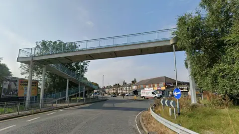 Google A roundabout under a pedestrian overpass. The sky is blue with light clouds. Trees and grass can be seen on the roundabout to the right. 