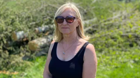 Fiona is wearing dark sunglasses and has blonde hair. She is stood in front of felled trees.