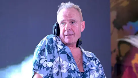 Getty Images Fatboy Slim, Norman Cook, stands on stage with one ear of his overhead headphones on. He is looking off to the side. He is an older man with grey, short hair and is wearing a light blue, dark blue and white hawaiian shirt.