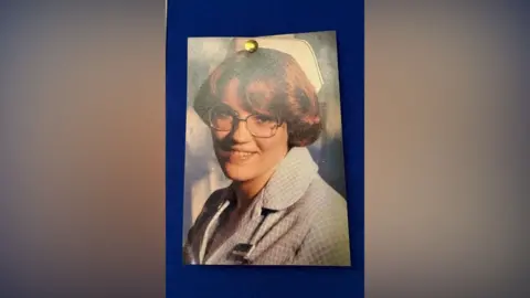 A photograph is pinned to a blue wall. It shows a young woman with short brown hair smiling into the camera. She is wearing big silver glasses and has a blue checkered shirt on and a white hat. 