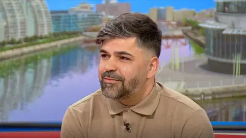 Sergio Aguiar sits on the BBC Breakfast sofa with a picture of Salford Quays behind him. He is wearing a beige polo shirt and has brown hair and facial hair.