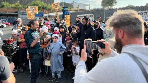 Aisha Iqbal/BBC A crowd of people, some of them children, many holding placards saying 'end the stink' and 'stop the fumes'. Some of the crowd wear face masks. A man stands in the middle holding a megaphone and addressing the crowd. Around them are are people filming on phones and cameras.