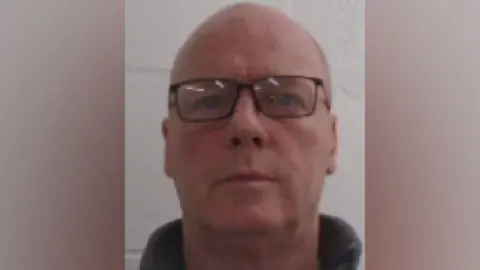 A mugshot photo of a man in his 50s wearing glasses with a shaven head.