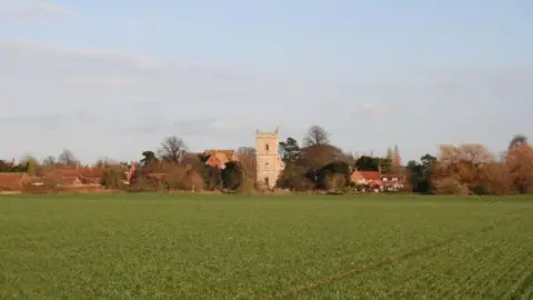 Richard Croft/Geograph A view across a green field of Bassingham village. A stone church tower stands in the centre, surrounded by red-brick properties and trees