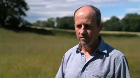 A man with a light blue shirt is standing on the right hand side of the photo. Grass is behind him and trees are in the distance.