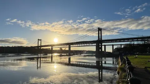 The Tamar Bridge on a sunny day with the sun reflecting in the water below it.