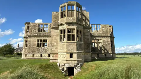 National Trust Images/Mike Selby Lyveden Lodge, a roofless late 16th Century building built out of cream-coloured stone. It was built in the shape of a cross and the view shows three of its four sides, with two sides on either side of a third which juts out in the middle. It is surrounded by grass and above is blue sky.
