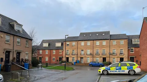 BBC A police car parked in a residential street, opposite a house with police tape blocking off the outside.
