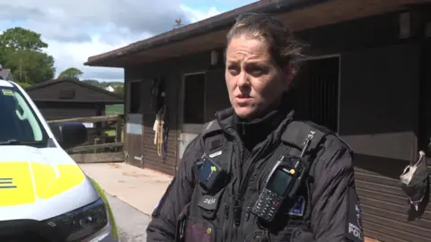 Lucy Wyatt looks toward the camera with a serious expression, wearing a police uniform and flanked by a police car and stables. 