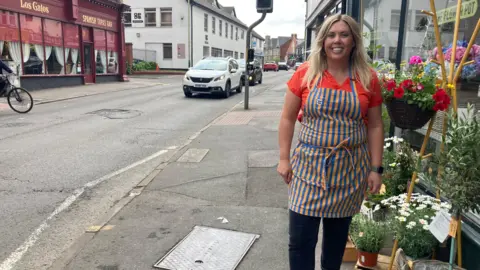 A smiling woman wearing a florist's apron standing outside a flower shop on Devizes Road in Swindon with a view down the road to the left.