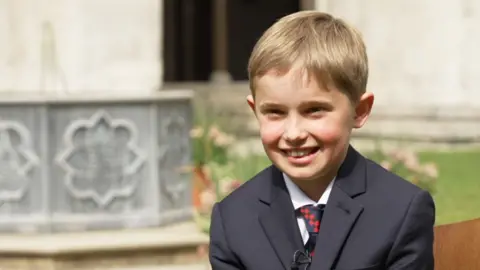 10-year-old Alexander Churchill wearing a suit and tie sat on a chair in a garden