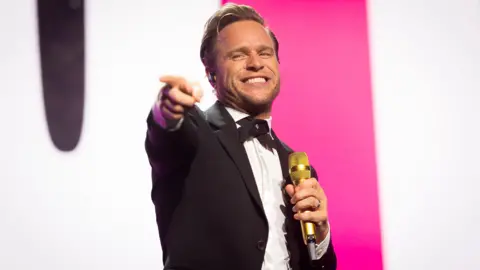 Olly Murs performs onstage - he is holding a grin microphone in one hand, grinning and pointing  with his other hand