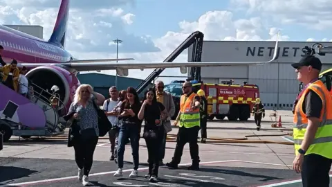 Passengers seen leaving the plane and walking towards the camera. It is a purple plane with WizzAir.com on the side. A fire engine is nearby with a fire fighter walking towards the plane, and two men in high vis clothing stand guiding passengers.