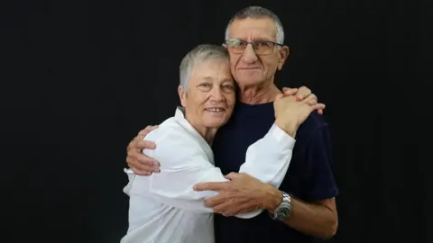 Magen Inon Bilha and Yakov Inon posing for a photograph against a black background. Bilha is wearing a white shirt and her husband is wearing a black t-shirt and he has glasses on.