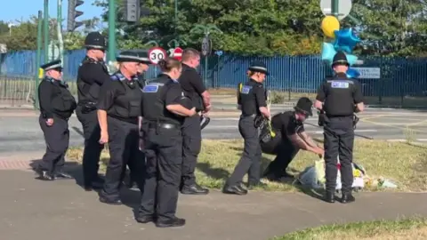 Eight police officers in uniform stand and watch as one of their colleagues puts down a floral tribute on an area of grass next to a road.