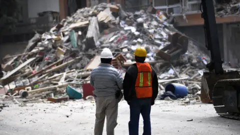 Getty Images Two men with their backs turned to camera survey a pile of rubble