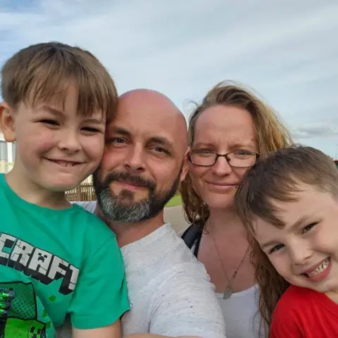 Tara Andrews The boy from the above picture wearing a green Minecraft t-shirt. He is being held by a bald man with a black/grey beard and a white t-shirt. Next to the man is a woman with neck-length dark blonde hair and a younger boy wearing a red t-shirt. All four are smiling at the camera.