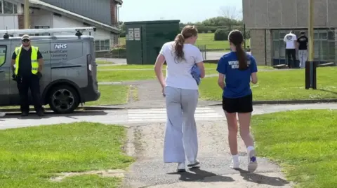 Two teenage girls walk down a path past an NHS van. One is wearing grey trousers and a white tshirt, the other is weawring a blue tshirt and black shorts. 
