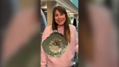 A woman in a pink jumper stands in an office. A top-down view of a volcano is superimposed in front of her