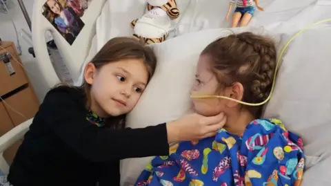 Supplied A girl tickling the chin of her sister in a hospital gown on a hospital bed