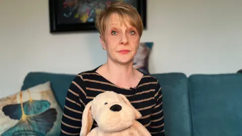 Caroline Dewson, a woman with short blonde hair, wearing a navy and camel-coloured stripey long-sleeved top. She is sitting on a teal-coloured sofa in her lounge and holding a sand-coloured stuffed toy dog.