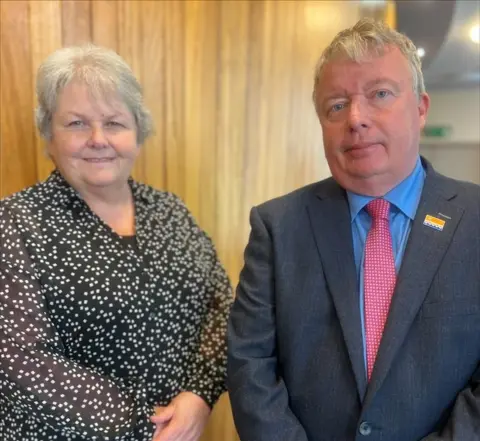A woman and a man smile at the camera. The woman has short white hair and is wearing a black and white blouse. The man has short grey hair and wears a blue suit, blue shirt and pink tie.