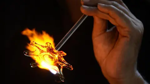 Close up of an artisan holding a glass straw object which opens up in a flower-like fashion near the bright yellow flame