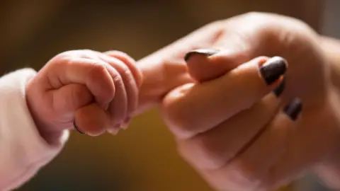 A baby's hand is gripping what appears to be a woman's index finger. The woman's finger nails are painted black or dark brown.