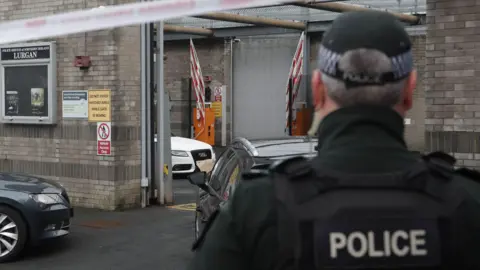 A uniformed police officer stands with his back to the camera outside Lurgan Police Station in the aftermath of an attempted bombing. There is a white and red police cordon tape above his head. Three vehicles are partially visible outside the station, which is a grey-bricked building. 