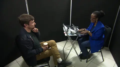 Dave Dean in a blue jacket and tan coloured trousers sat cross legged on a chair opposite a woman in a blue jacket who is sat on a chair and typing on a laptop. The pair are in a room surrounded by black walls