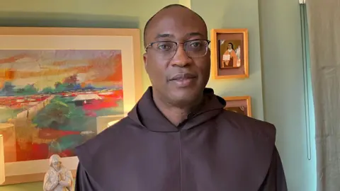 Father Alexander Ezechukwu in a traditional brown robe. He has a shaved head and glasses. behind him is a green wall with a colourful painting hung on it.