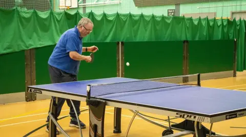 An older man prepares to hit a ball with a small paddle on a blue table tennis table in a sports hall. There are green nets behind him and court markings on the floor.