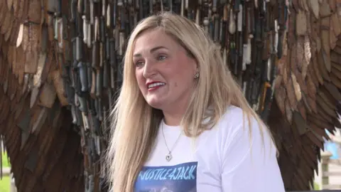 Head and shoulder shot of Zoe McGill, a woman with long blonde hair and a serious expression. She is wearing a white T-shirt with a "Justice 4 Jack" logo, and standing in front of a metal sculpture made of knives.