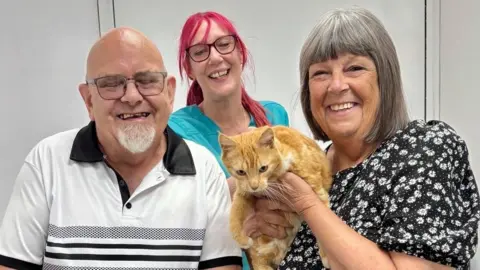 Robert Poile, in a black and white top, and Debbie Poile, wearing a black and white dress, are at the vets holding their ginger cat Primrose, with veterinary nurse Chris Guscott behind them.
