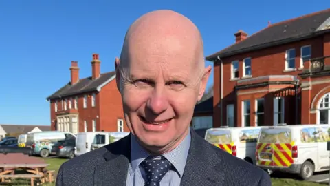 Tegryn Jones, a man wearing a grey and navy blue suit, stands in front of red brick houses looking at the camera