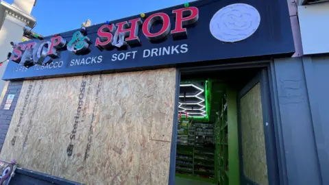 Martin Giles/BBC Shop windows boarded up. The entrance is open and neon lights are on inside. Vapes and cigarettes are on display at the back of the shop.